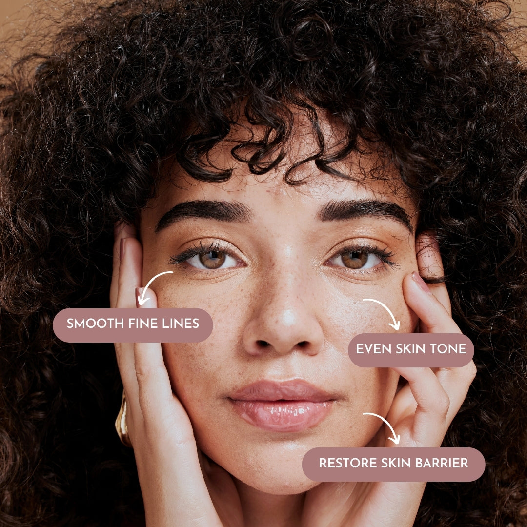 Close-up of a woman of color holding her face and looking straight with three pink labels on the product's skin effect on her face. On the left, under her eyes, are "smooth fine lines," and on the right are "even skin tone" and "restore skin barrier". 
